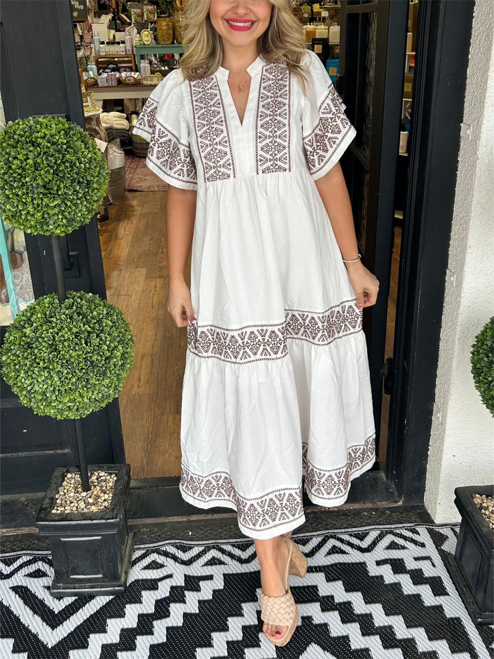 Woman in white boho V-neck midi dress with brown embroidered patterns, short sleeves, and tiered skirt.
