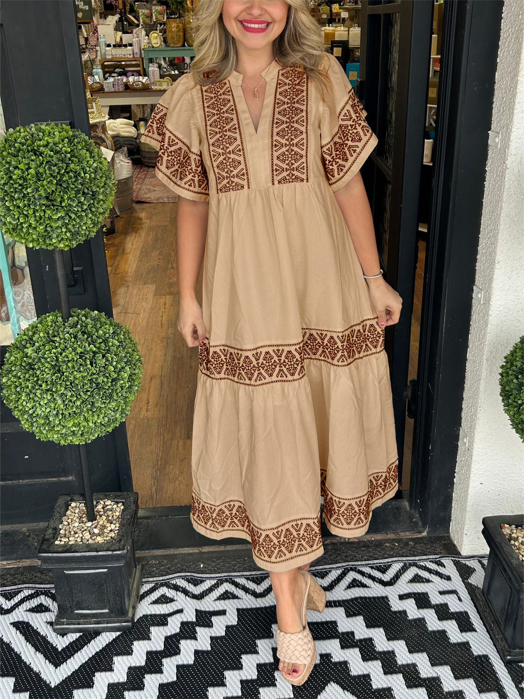 Woman in a beige Abigail boho V-neck midi dress with brown embroidered patterns, standing in a doorway.