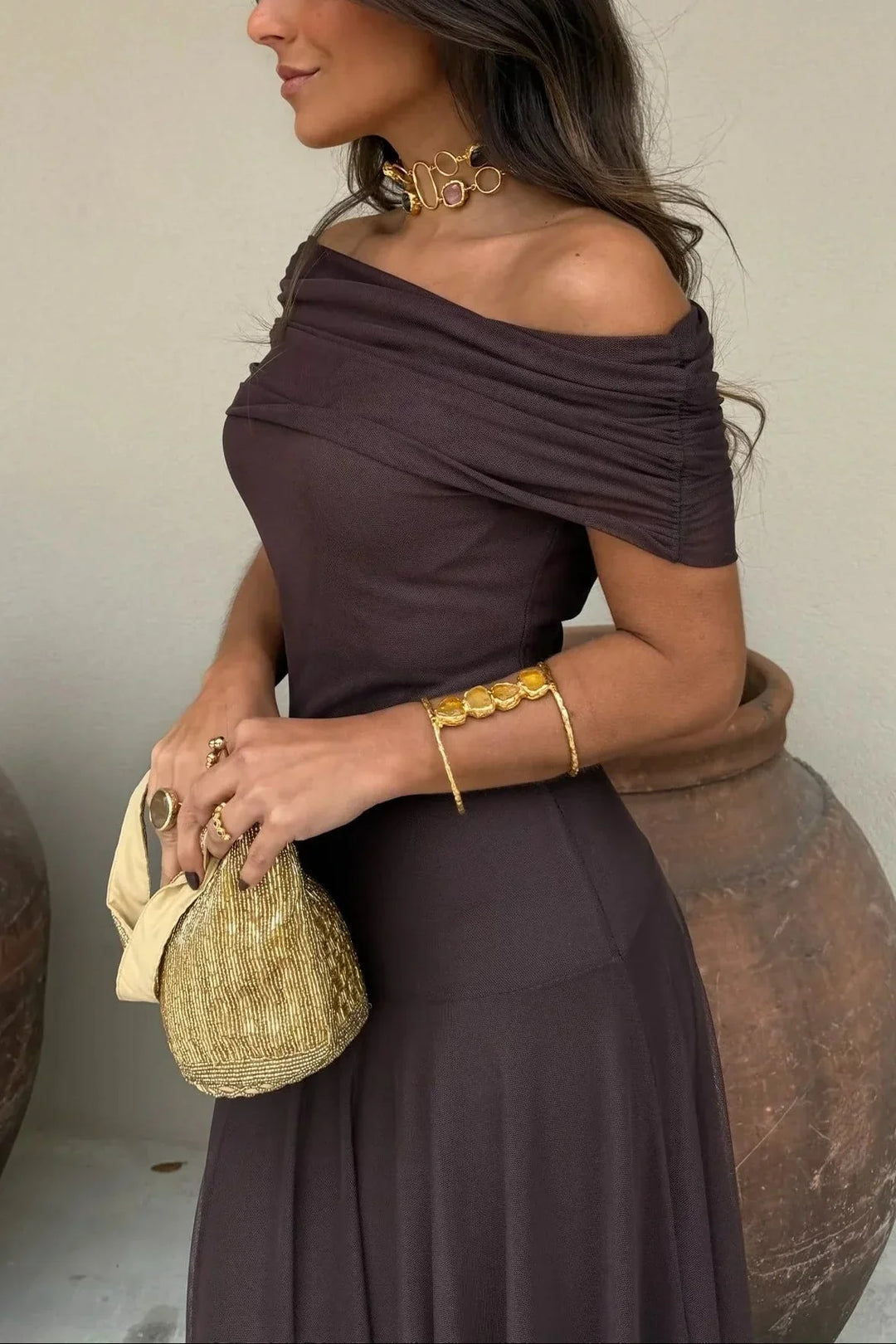 Woman in a dark brown one-shoulder maxi dress with draped bodice, holding a gold beaded clutch.