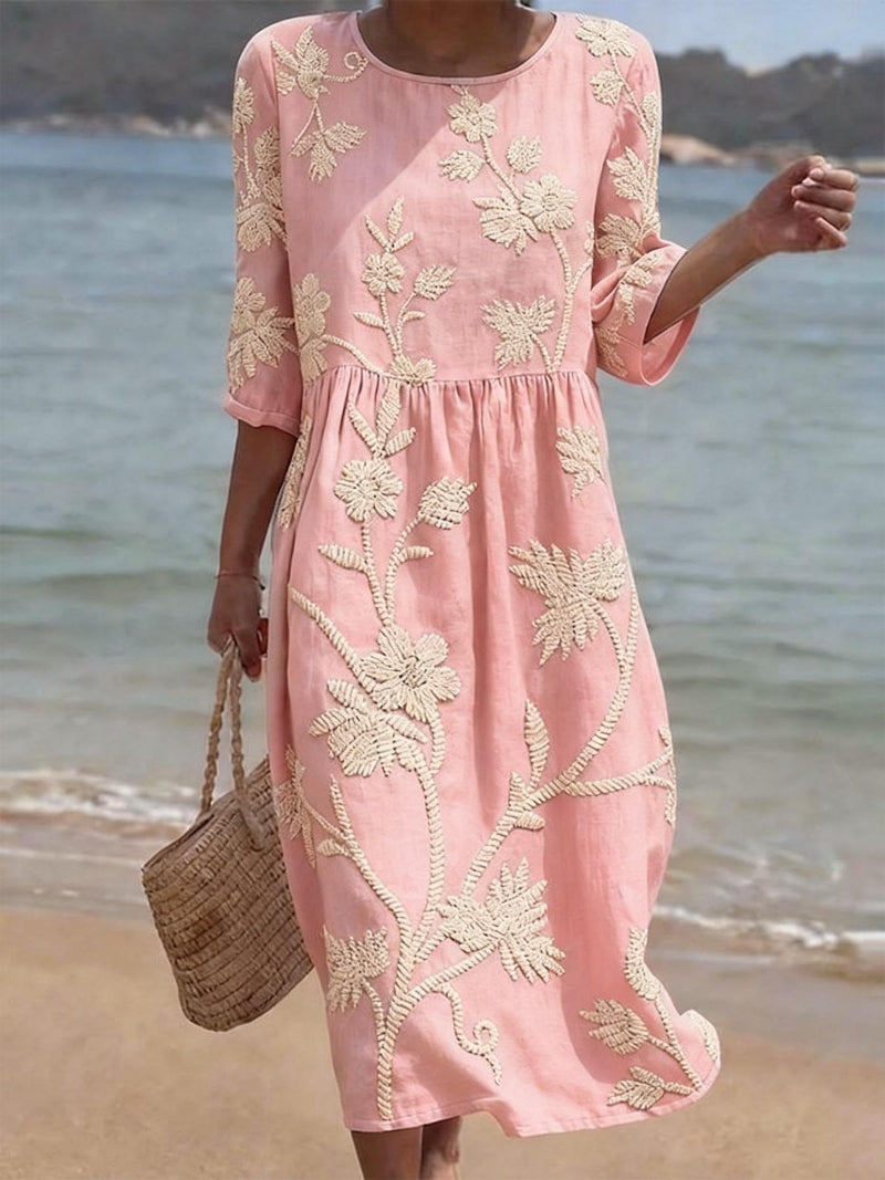 Women's pink midi dress with cream floral embroidery, three-quarter sleeves, and a relaxed fit, worn on a beach.