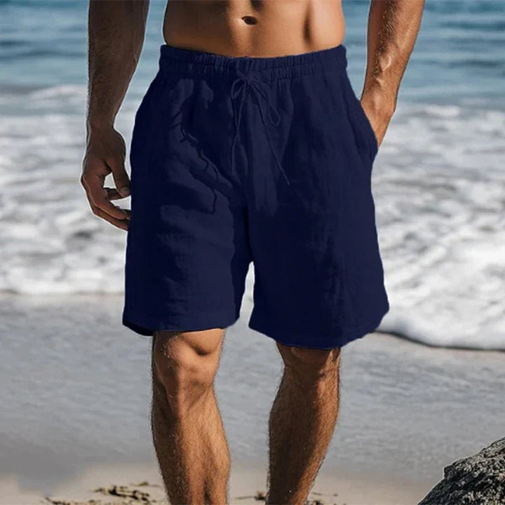 Men's navy linen drawstring shorts with side pockets, worn on a beach.