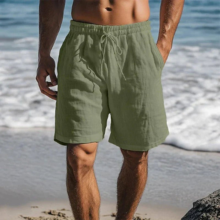 Men's olive green linen drawstring shorts with side pockets, shown on a beach.
