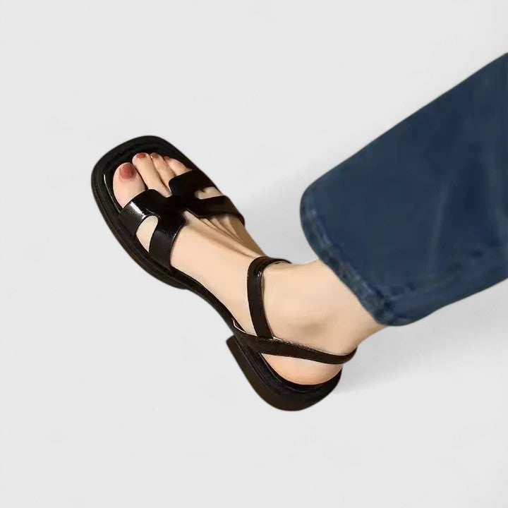 Black leather ankle strap flat sandals with open toe and gold buckle.