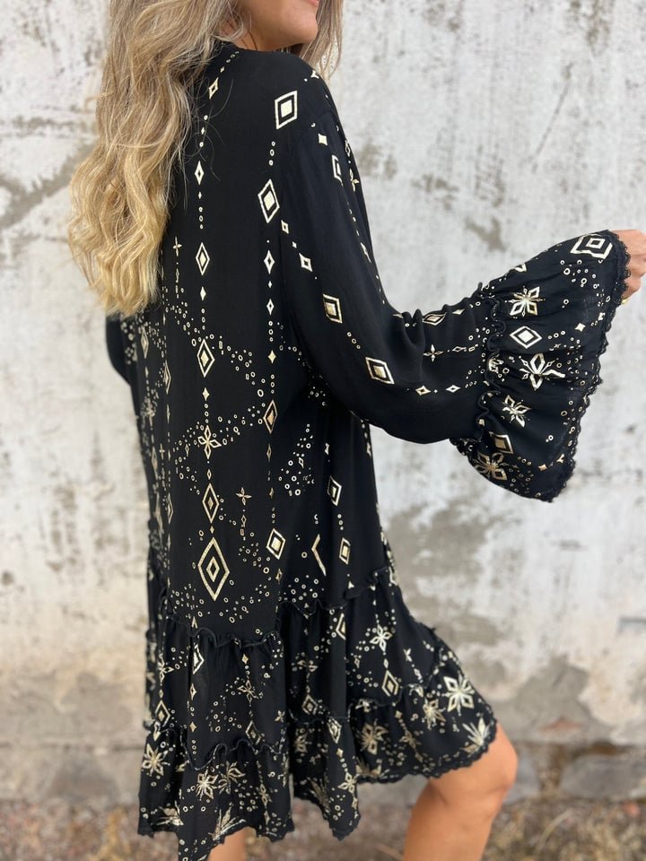 Black tiered boho V-neck dress with gold geometric and floral patterns, three-quarter bell sleeves, and ruffled hem.