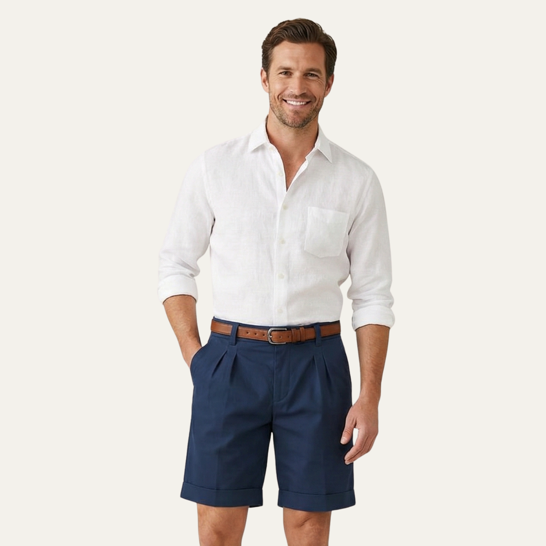 Man wearing navy Julian pleated chino shorts with cuffed hems, a brown belt, and a white linen shirt.