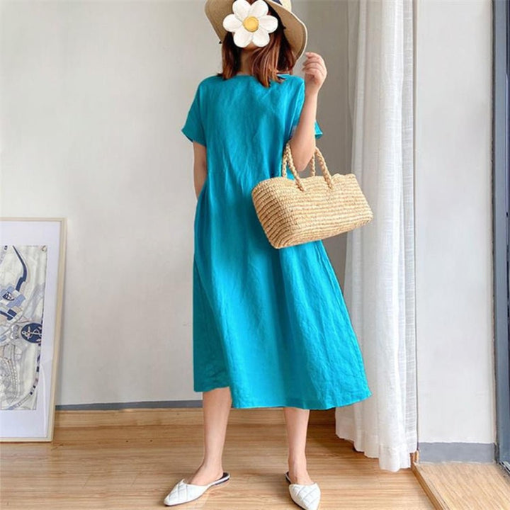 Woman in a blue linen short sleeve midi dress, straw hat, and woven bag.
