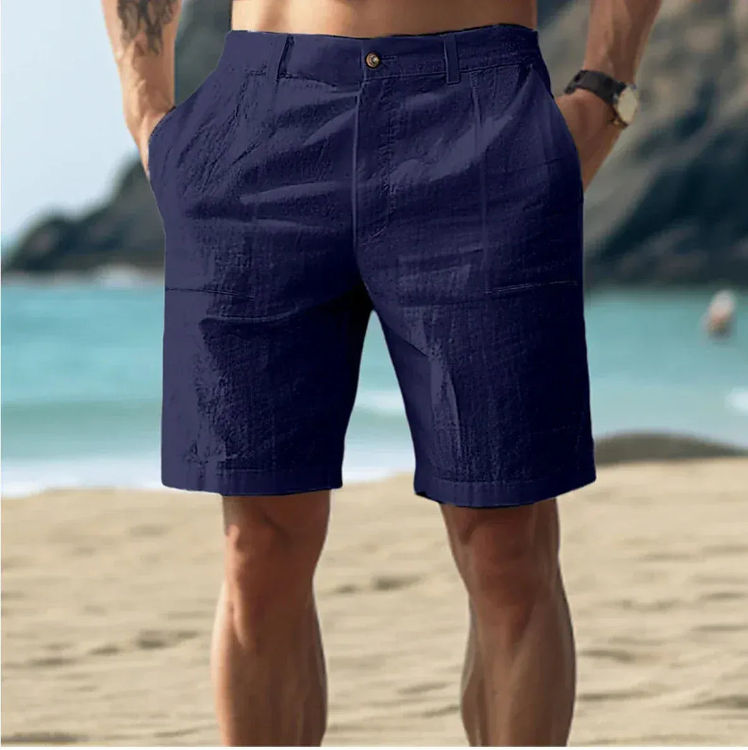 Men's Nico above-knee button-front shorts in navy blue, shown on a person standing on a beach.