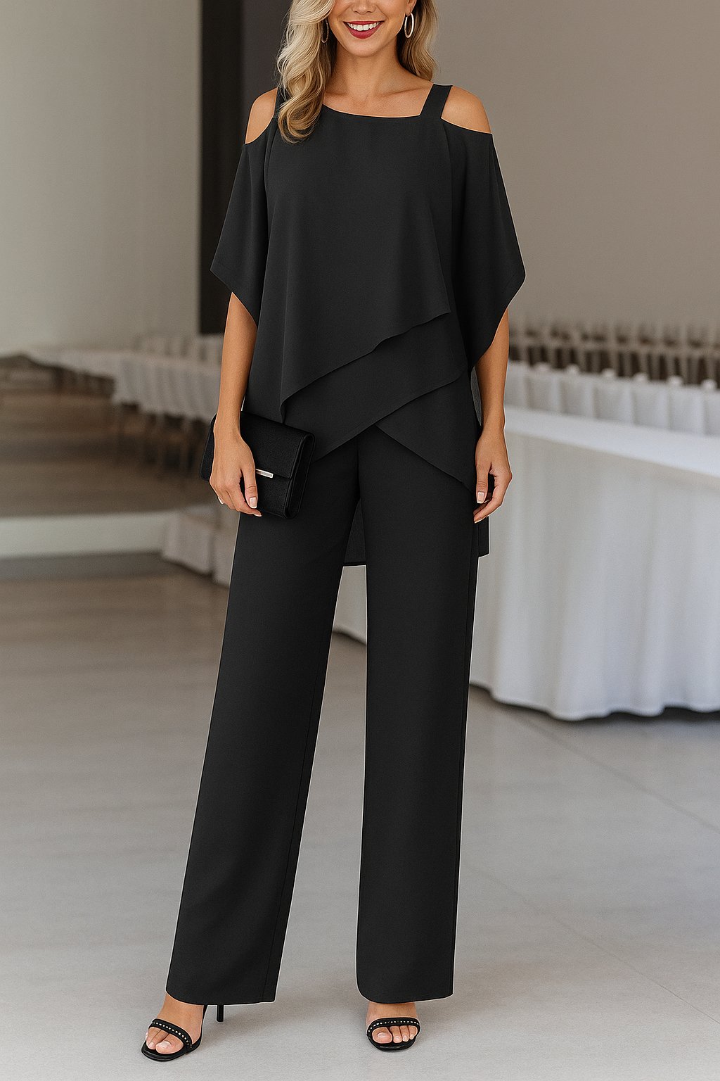 Woman in a black cold shoulder wide-leg jumpsuit with a draped overlay, holding a clutch.