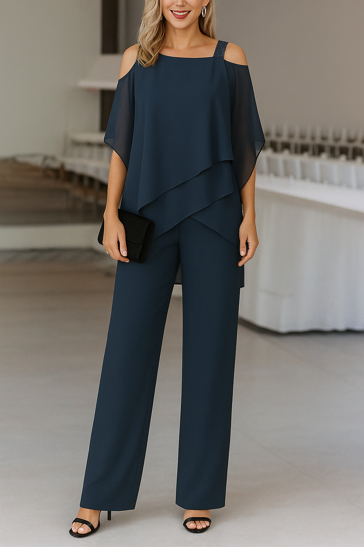 Woman in a dark teal cold shoulder wide-leg jumpsuit with a layered draped overlay, holding a black clutch.